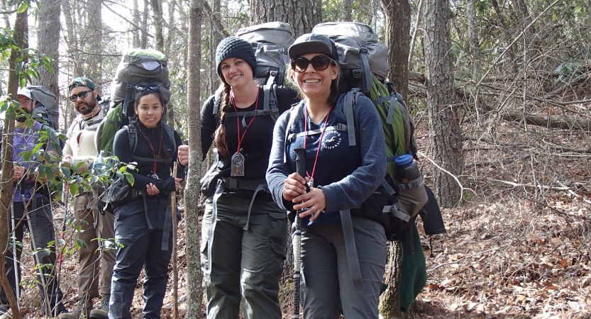 A group of people wearing backpacks smile in a wooded landscape. 